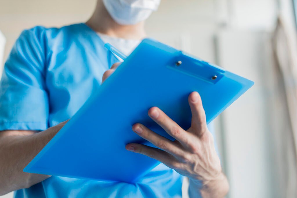 A imagem mostra o tronco de um homem que veste uma roupa azul, que é um uniforme usado por profissionais de Enfermagem. Nas mãos dele há uma prancheta azul, na qual ele faz anotações. 