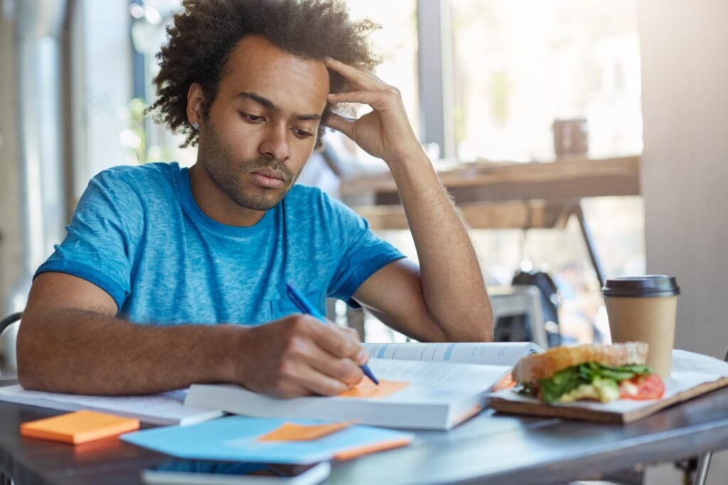 A imagem mostra um rapaz negro de cabelo crespo. Ele está sentado em frente a uma mesa de estudos e faz anotações em um papel adesivo laranja, que está colado em um livro de estudos.  