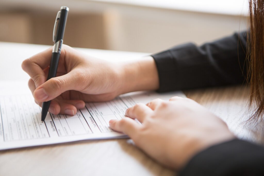 A imagem mostra as mãos de uma mulher branca sobre uma mesa de madeira. Ela está preenchendo um formulário com uma caneta preta. 