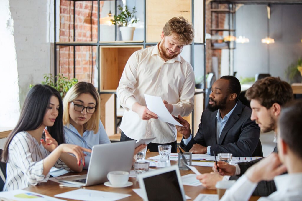 A imagem mostra um grupo de pessoas, duas mulheres e quatro homens, reunidos em torno de uma mesa de escritório. Eles conversam e parecem resolver problemas com o trabalho. 