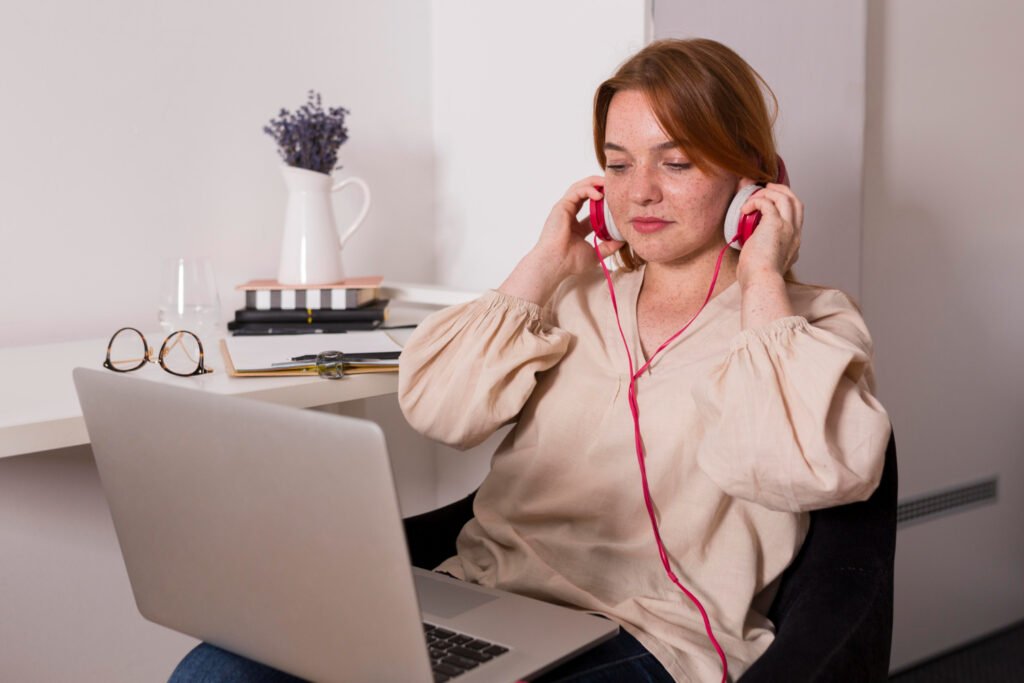 La imagen muestra a una mujer blanca pelirroja sentada en un sillón. Sobre sus piernas tiene una computadora portátil a la cual está mirando. Con ambas manos, sostiene sus audífonos rosas a la altura de sus orejas. 