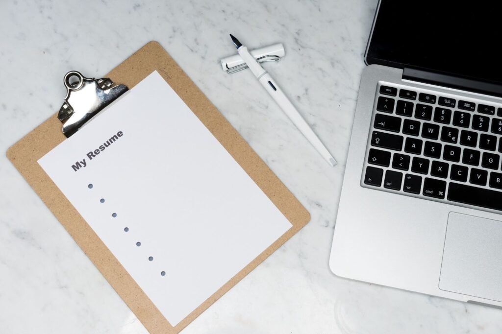 A imagem mostra uma mesa de mármore onde estão uma prancheta, uma caneta branca e um laptop prata com teclas pretas. Na prancheta, há um papel onde está escrito “my resume”, e alguns tópicos em um esquema de lista. 