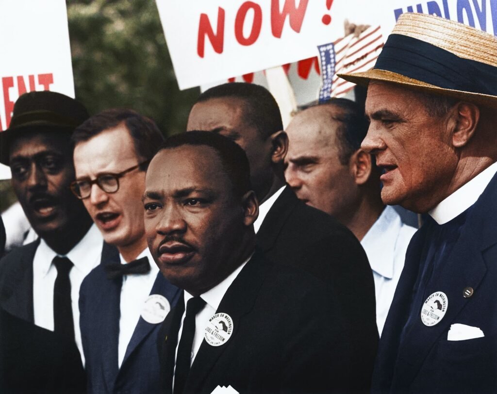  Na imagem, é possível ver seis homens, com a figura de Martin Luther King Jr. em foco. A maioria dos presentes estão vestidos de terno e, a julgar pelos bottons usados e pelas placas de protesto parcialmente expostas, pode-se deduzir que se trata de um cenário de manifestação e protestos. 