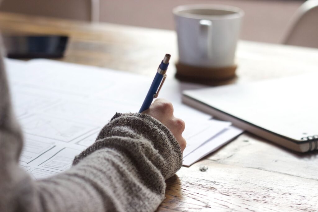 A imagem mostra uma pessoa de blusa de manga longa cinza. Ela está escrevendo com uma caneta azul em papéis espalhados sobre uma mesa de madeira. Ao fundo, é possível ver um caderno, uma xícara de café e um aparelho smartphone. 