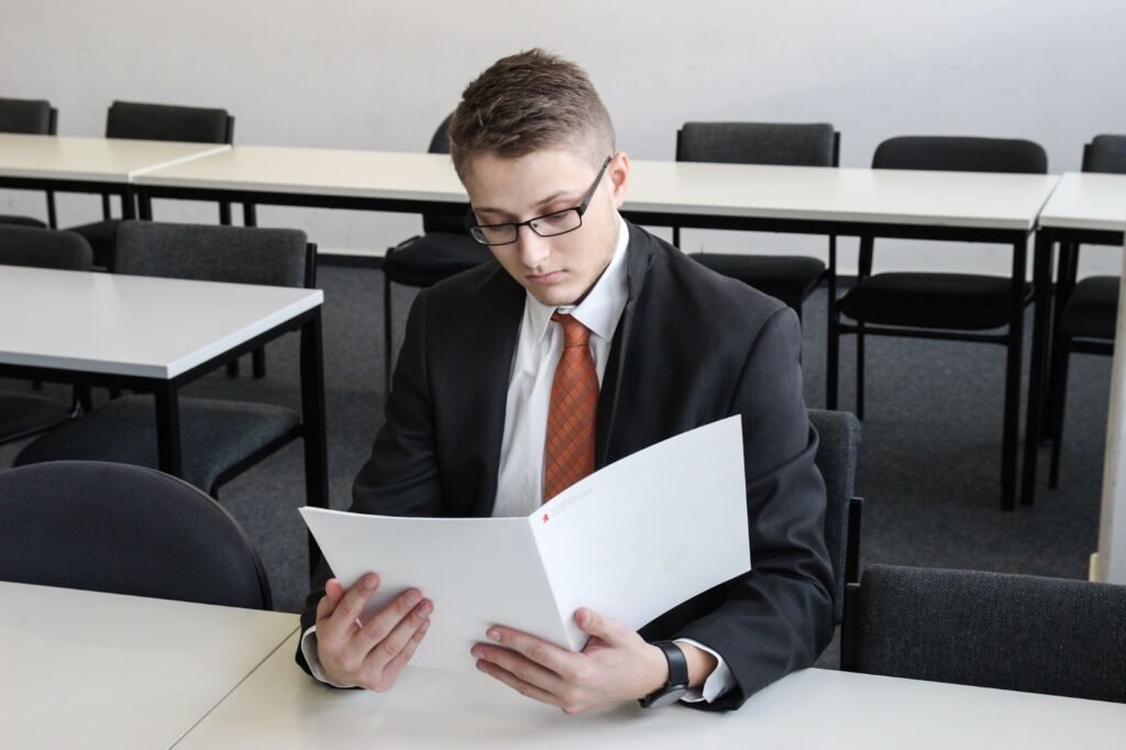 A imagem mostra um homem loiro de óculos quadrados pretos. Ele veste um terno com uma gravata laranja e está sentado atrás de uma mesa. Com as mãos, ele segura um documento aberto.