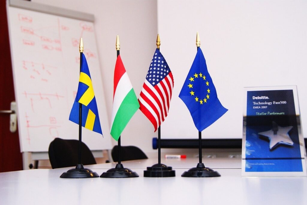 La imagen muestra cuatro minibanderas ondeando en un soporte de plástico negro. De izquierda a derecha, está la bandera de Suecia, la de Hungría, la de Estados Unidos y la de la Unión Europea. Junto a ellas, hay una pequeña placa azul que tiene escrito “Deloitte, Technology Fast500 emea 2007. Stellar Performers”. Todo está sobre una mesa blanca. Al fondo de la imagen, se puede ver un tablero blanco y dos sillas negras que están junto a la mesa. 