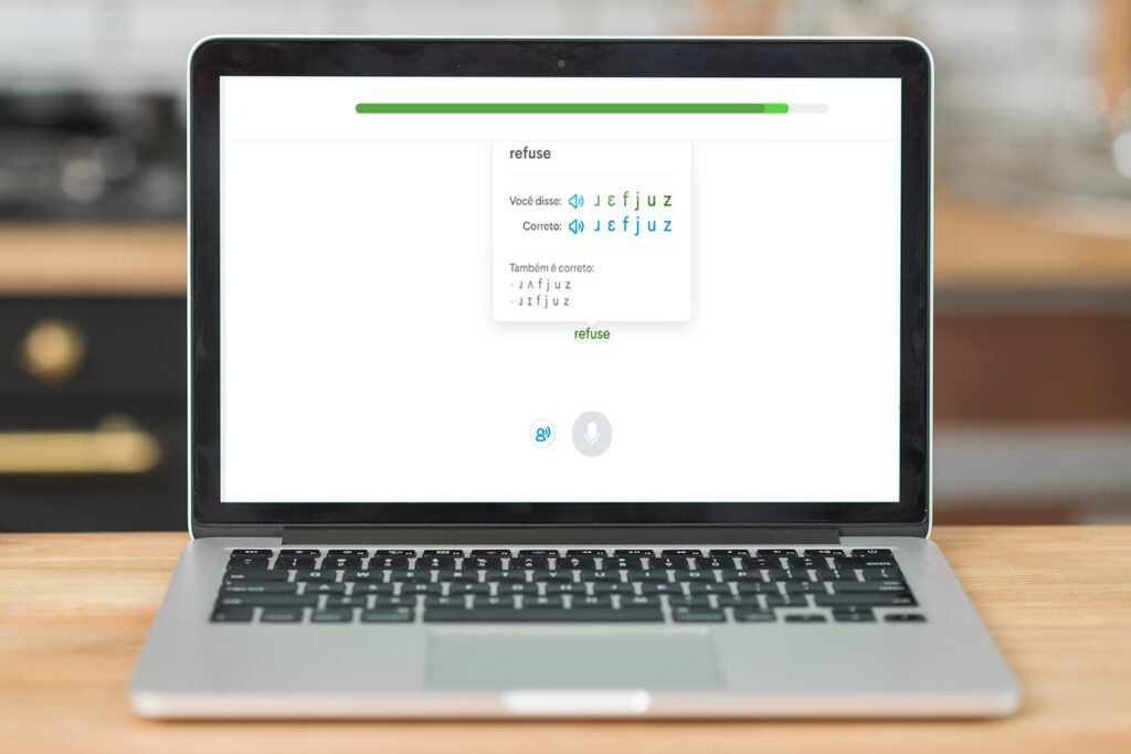 A imagem mostra um laptop prata, que está aberto sobre uma mesa de madeira. Na tela dele, há em destaque a página de exercício da Slang, na qual é possível ver a palavra “refuse”. 