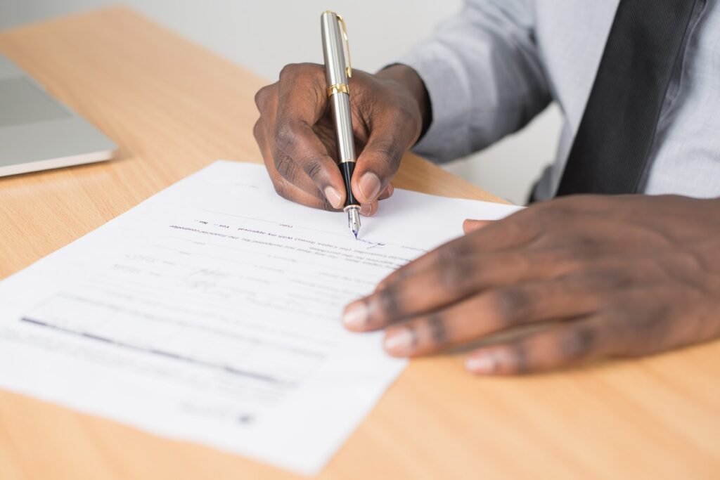 A imagem mostra as mãos de um homem negro sobre uma mesa de madeira clara. Com a mão direita ele segura uma caneta e preenche um papel branco que também está na mesa. A mão esquerda dele está apoiada nessa folha.