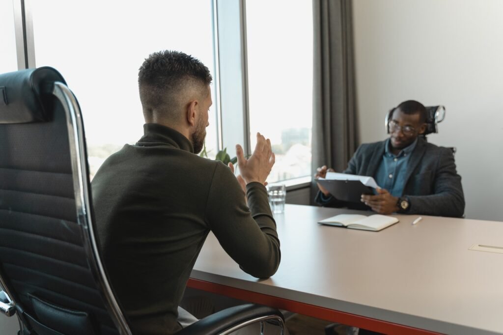 La imagen muestra a dos hombres negros en una sala de reuniones. Están de frente y separados por una mesa gris. En la parte izquierda de la foto, hay un hombre joven que tiene el pelo rizado en la parte superior de la cabeza y los lados rapados. Frente a él, hay un hombre negro que parece ser un poco mayor. Lleva un traje, gafas y está tomando notas en una hoja apoyada en un portapapeles que tiene en la mano izquierda.