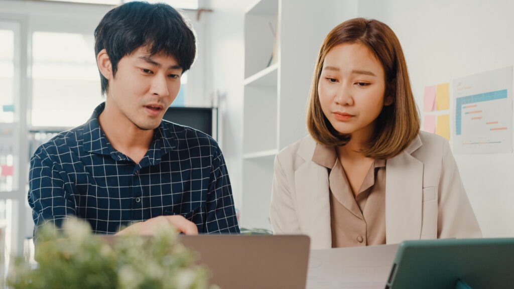 A imagem mostra um rapaz asiático e uma mulher asiática lado a lado. Eles estão olhando para a tela de um notebook, que aparece desfocado.