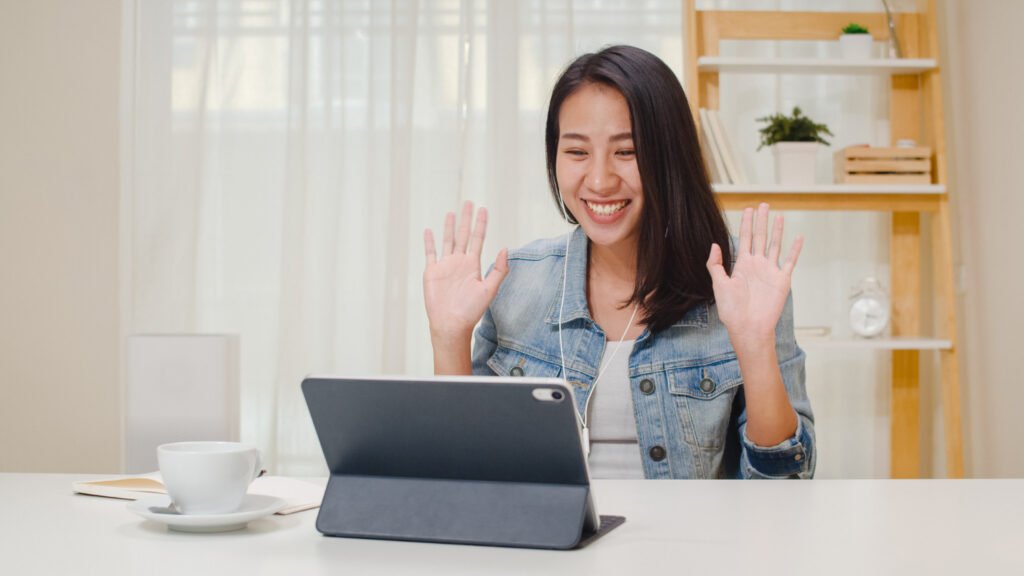 A imagem mostra uma mulher asiática, de pele branca e cabelo castanho escuro liso na altura do ombro, em frente a uma mesa. Ela está sorrindo, suas mãos estão um pouco levantadas e abertas na altura dos ombros, como se estivesse comemorando algo. Ela olha para um tablet, que está apoiado em uma capa cinza em cima da mesa. 