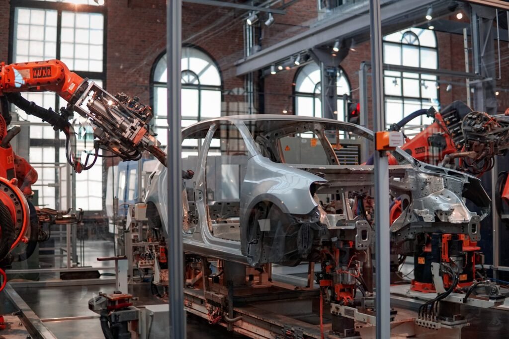 A imagem mostra a carcaça de um carro prata sustentada por uma máquina laranja. Na foto, o carro parece estar em processo de montagem, ainda na fábrica do setor automotivo. 