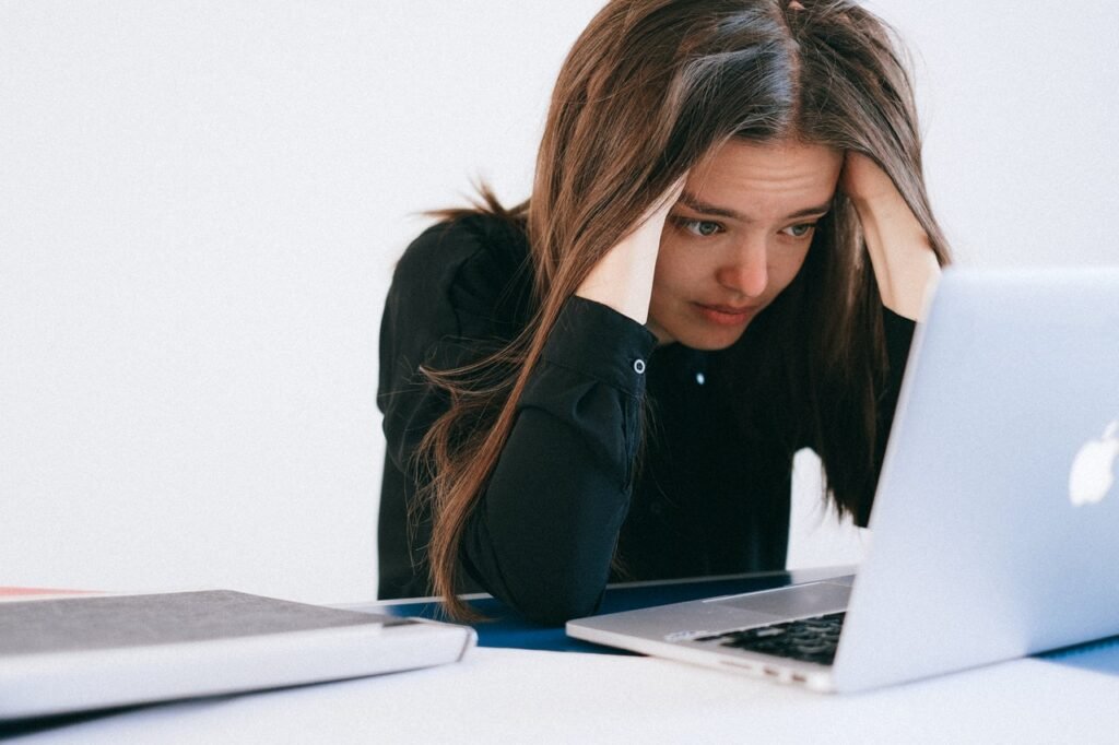 Uma mulher branca de cabelos castanhos lisos está em frente a um laptop prata. Ela olha para a tela e segura a cabeça com as mãos, como se estivesse preocupada ou insatisfeita, um sinal da falta de reconhecimento profissional nas empresas. 