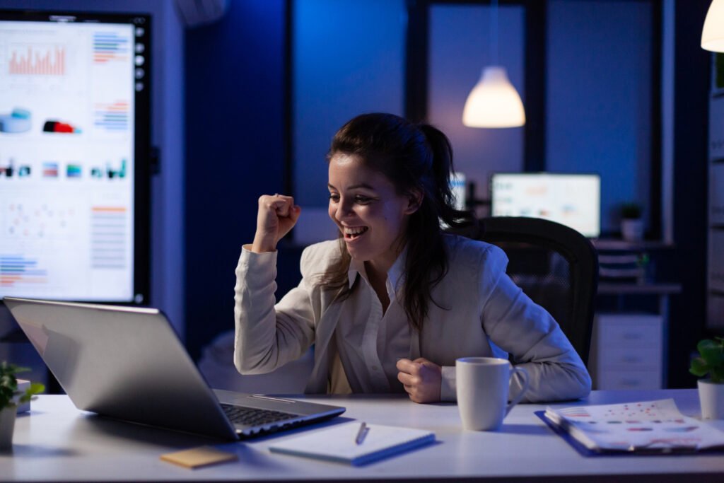  Uma mulher branca de cabelo castanho aparece em frente a um laptop prata. Ela está sorrindo e faz um gesto com as mãos, uma espécie de soco no ar, como se estivesse comemorando algo. 