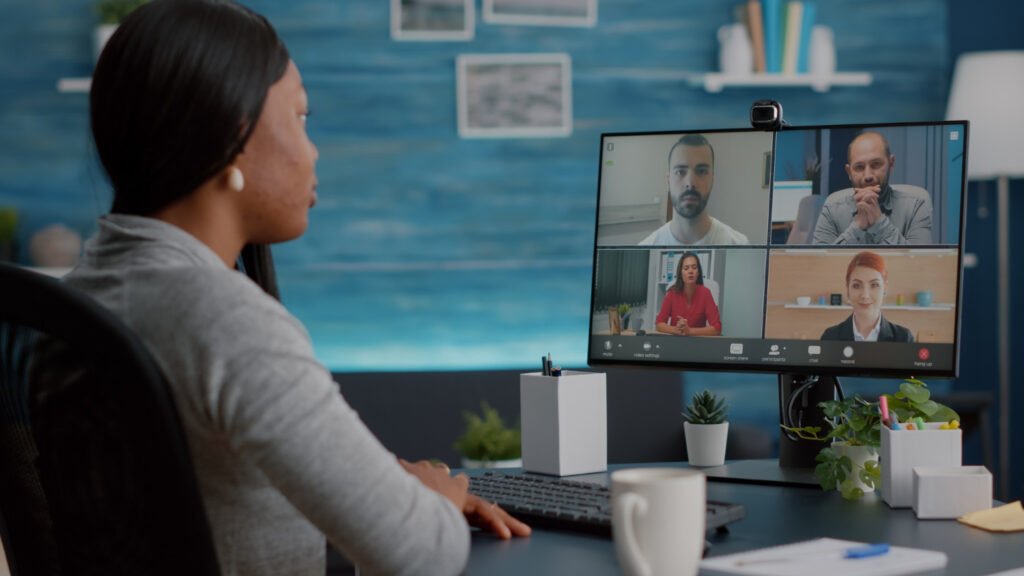 A imagem mostra uma jovem negra, de cabelo preto liso sentada em frente a uma mesa, na qual há o monitor de um computador. Na tela é possível ver uma videochamada com mais quatro pessoas, da esquerda para a direita, há dois homens brancos, uma mulher branca de cabelo castanho e uma mulher branca ruiva.