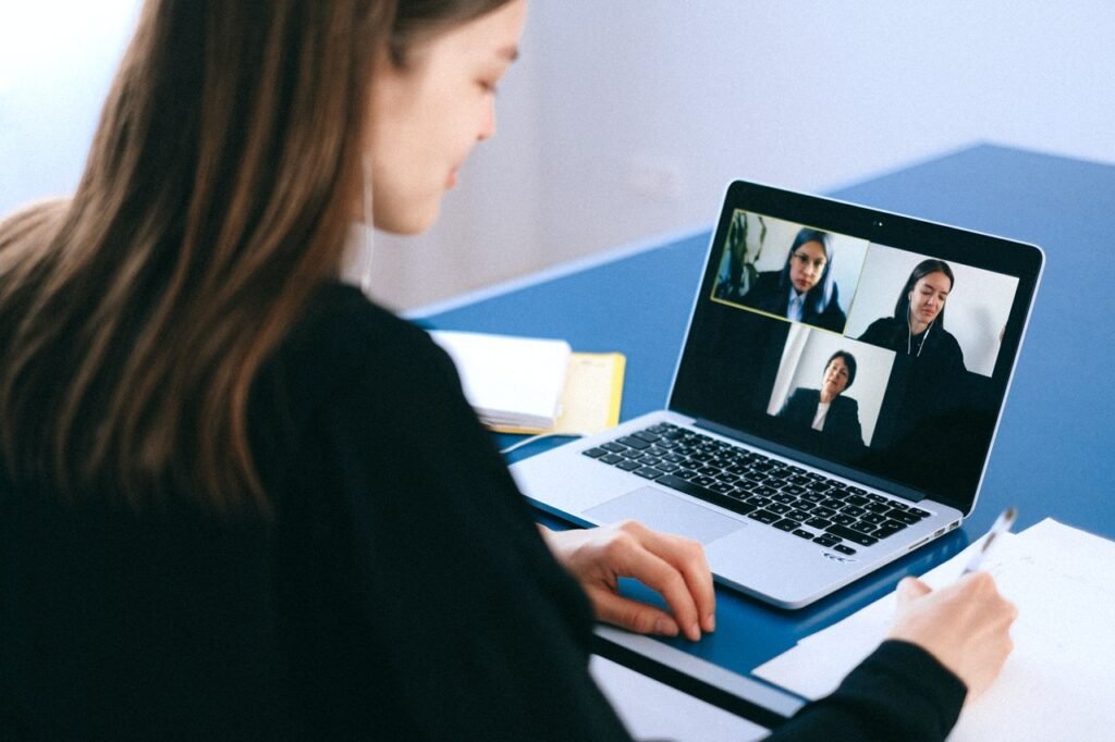 A imagem mostra uma mulher branca em frente a um laptop prata, que está em cima de uma mesa azul. Em sua tela é possível ver uma videoconferência com outras três mulheres.