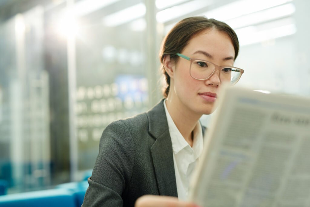 A imagem mostra uma mulher asiática de pele branca e cabelo castanho-claro liso. Ela usa óculos quadrados em tom marrom-claro. A mulher olha para um jornal que está à sua frente e aparece desfocado na foto. 