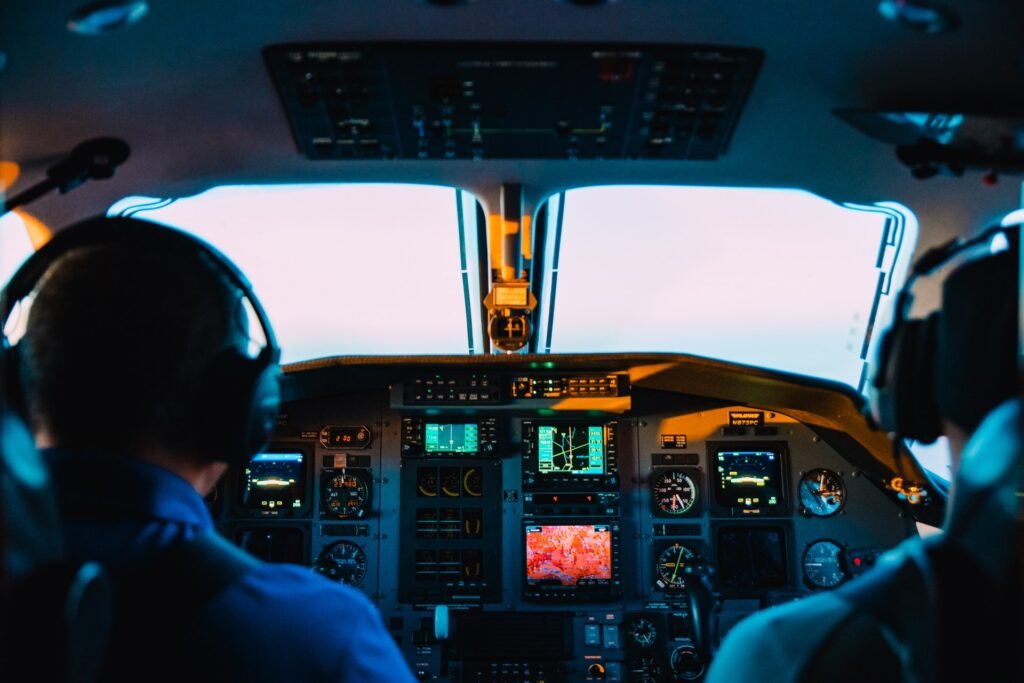 Dois pilotos de avião aparecem de costas para a imagem. Eles estão em uma cabine de comando da aeronave e representam como se tornar piloto de avião de companhias aéreas.  