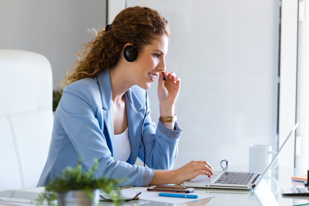 Uma mulher branca, de cabelo cacheado castanho-claro está em frente a uma mesa de escritório. Ela olha e mexe em um laptop prata, como se estivesse atendendo a um cliente por chamada de voz. 