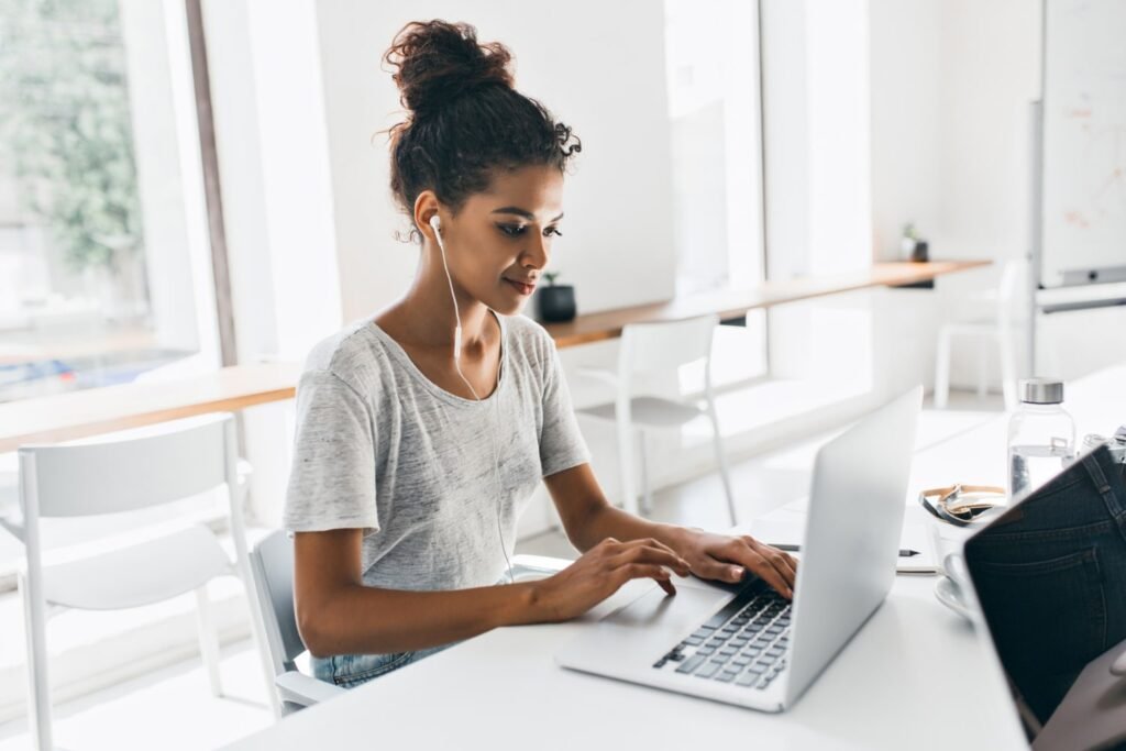 Uma mulher negra, de cabelo cacheado castanho-escuro em um coque, está em frente a uma mesa de trabalho branca. Ela mexe em um laptop prata. 