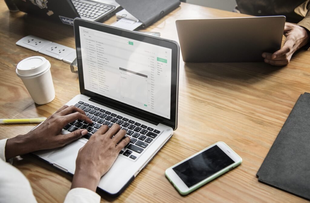 La imagen muestra las manos de una persona negra sobre una laptop plateada. La pantalla muestra la bandeja de entrada de un correo electrónico y la persona parece estar escribiendo un mensaje nuevo.