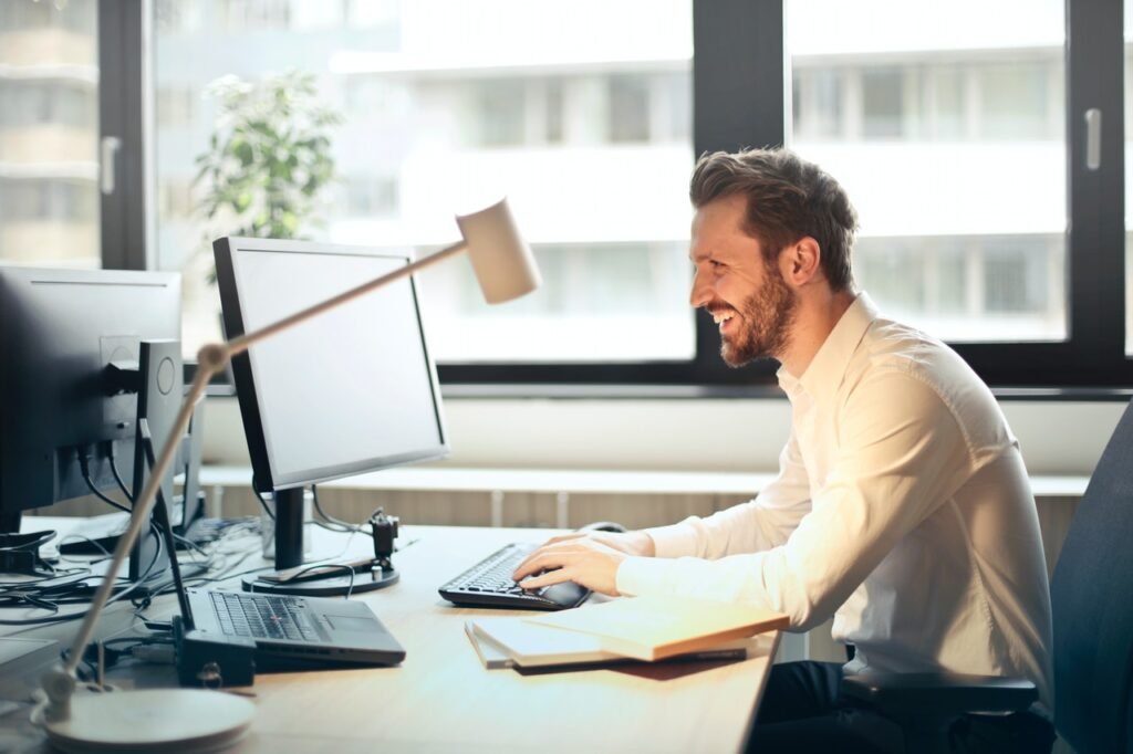 La imagen muestra a un hombre blanco sentado en un escritorio de madera. Él se ubica del lado derecho de la imagen y se ve de perfil. Frente a él, hay un monitor y una laptop, la cual está mirando.