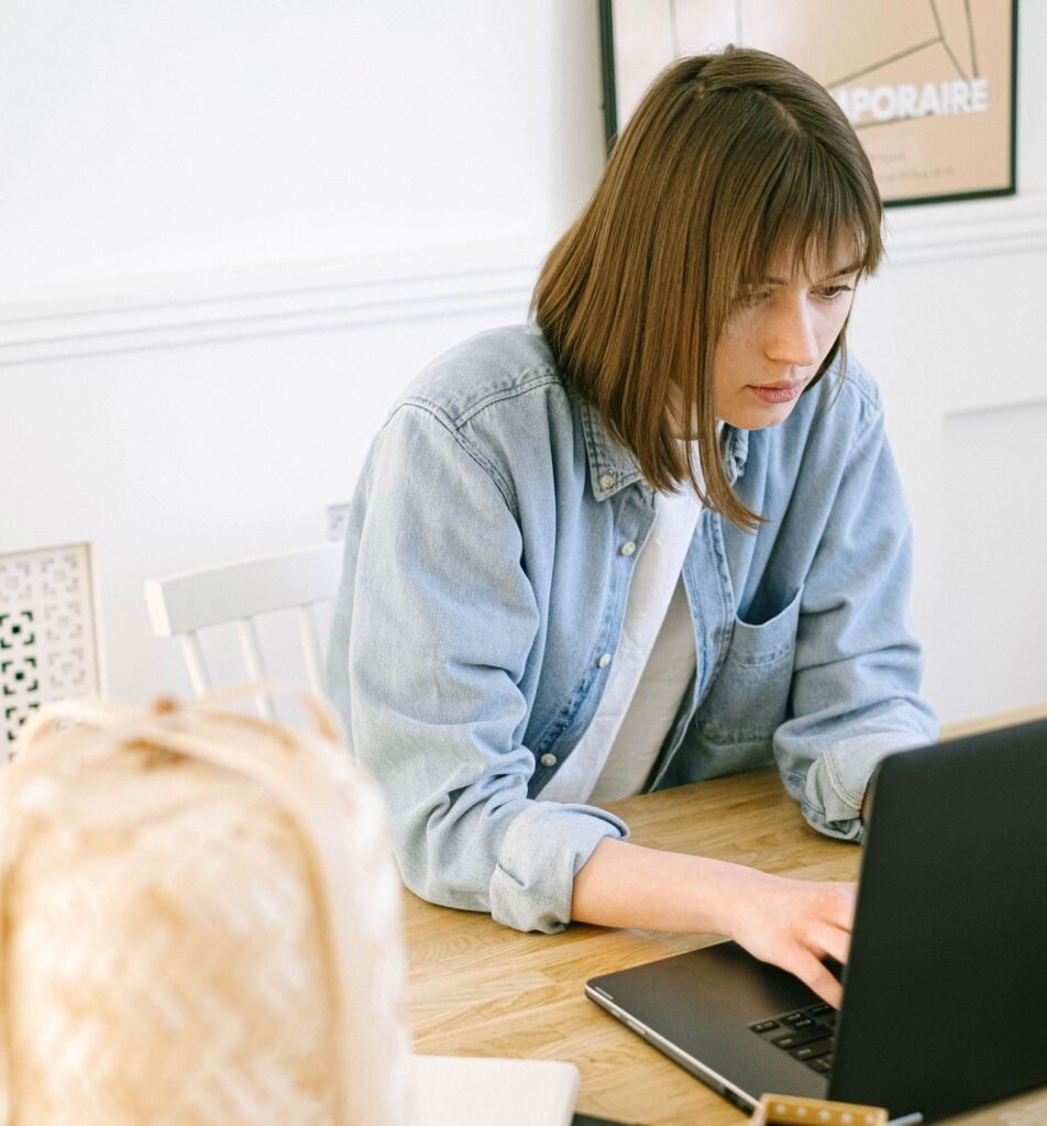  La imagen muestra a una mujer blanca de cabello liso, castaño claro y que está sentada en un escritorio. Ella tiene las manos apoyadas sobre el teclado de una laptop negra, la cual está observando. 