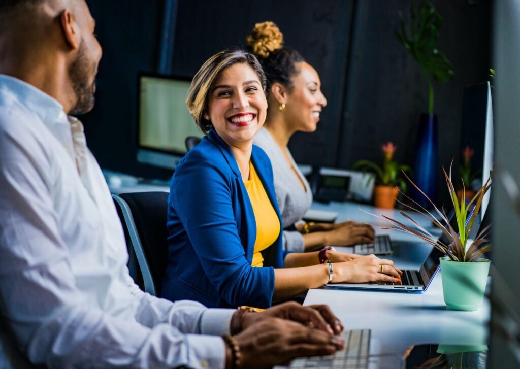 La imagen muestra a un hombre y dos mujeres frente a una mesa de oficina con computadoras sobre ella. Destaca en la imagen una mujer blanca de pelo castaño con mechas rubias, quien sonríe y mira al hombre que está a su lado y  se observa de perfil en la imagen.