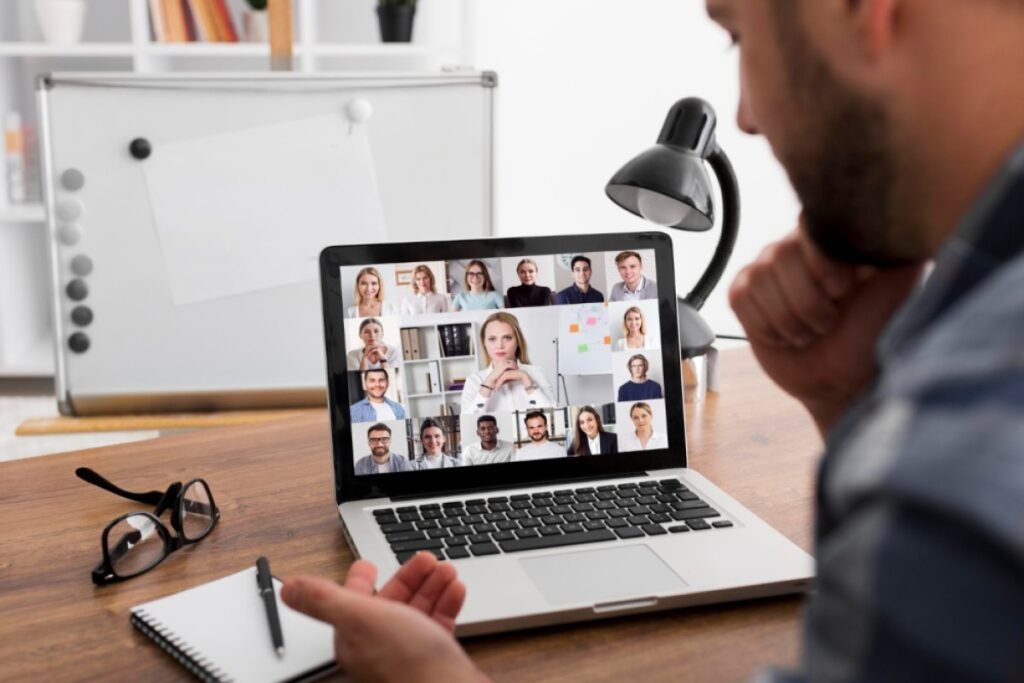 Um homem branco está olhando para um laptop prata. Na tela é possível ver uma videochamada com diversas pessoas. 