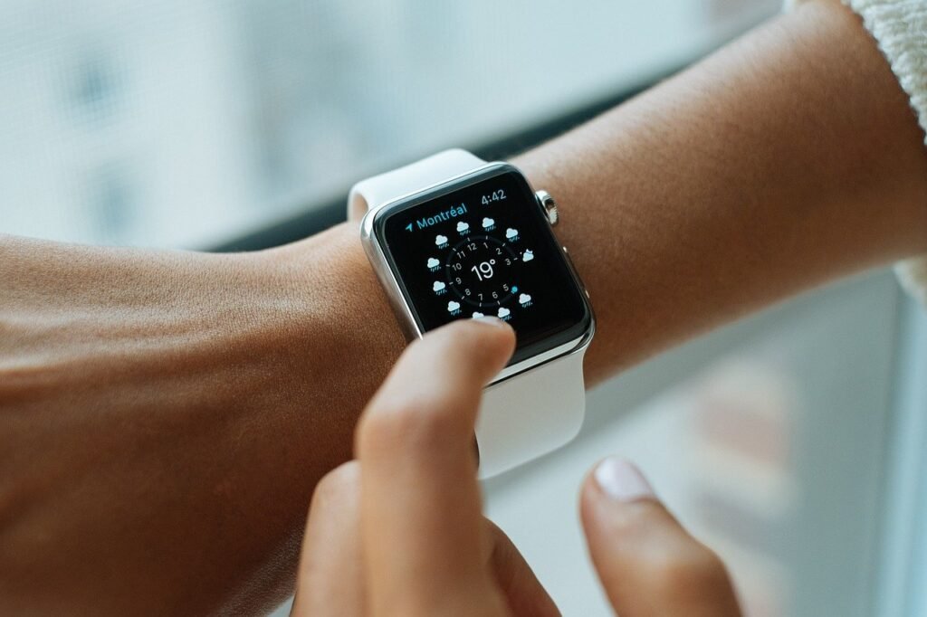 O braço esquerdo de uma pessoa negra de traços finos aparece na imagem. Nele é possível ver um smartwatch branco que mostra a previsão do tempo. 