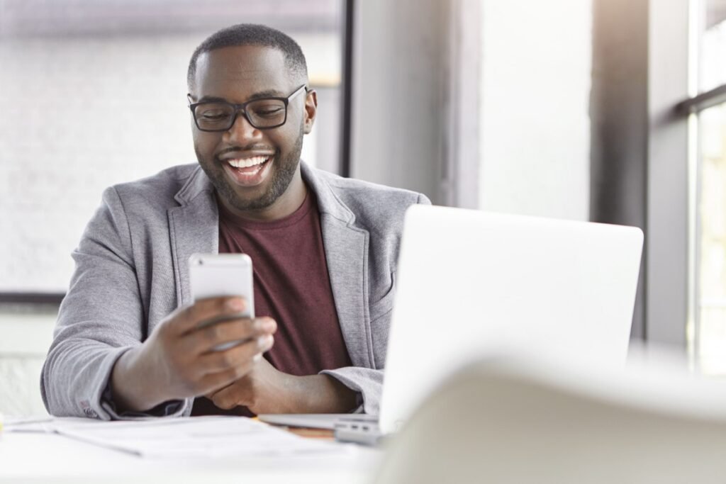  La imagen muestra a un hombre negro sentado frente a un escritorio. Usa gafas negras cuadradas, una camiseta roja y un blazer gris. En su mano izquierda, sostiene un teléfono móvil plateado. Su mirada está puesta en la pantalla del celular y sonríe. En la imagen también se aprecia un poco una laptop y algunos papeles sobre la mesa. 