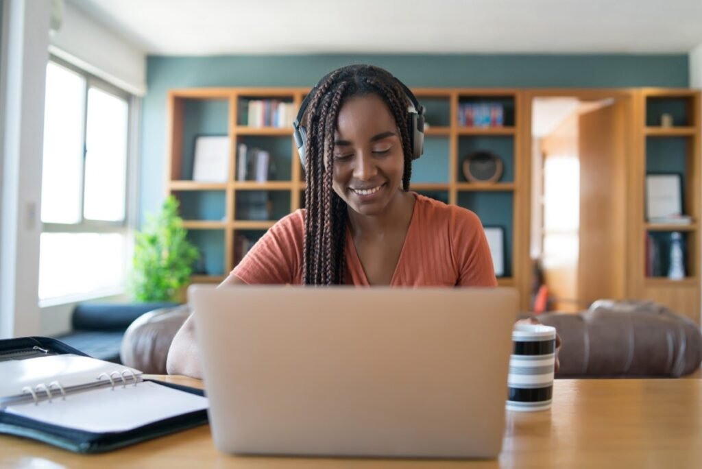 La imagen muestra a una mujer negra frente a una laptop plateada que está sobre una mesa de madera. Tiene el pelo trenzado, lleva un headset, mira la pantalla del ordenador y sonríe. En el fondo, se puede ver un librero de madera. 