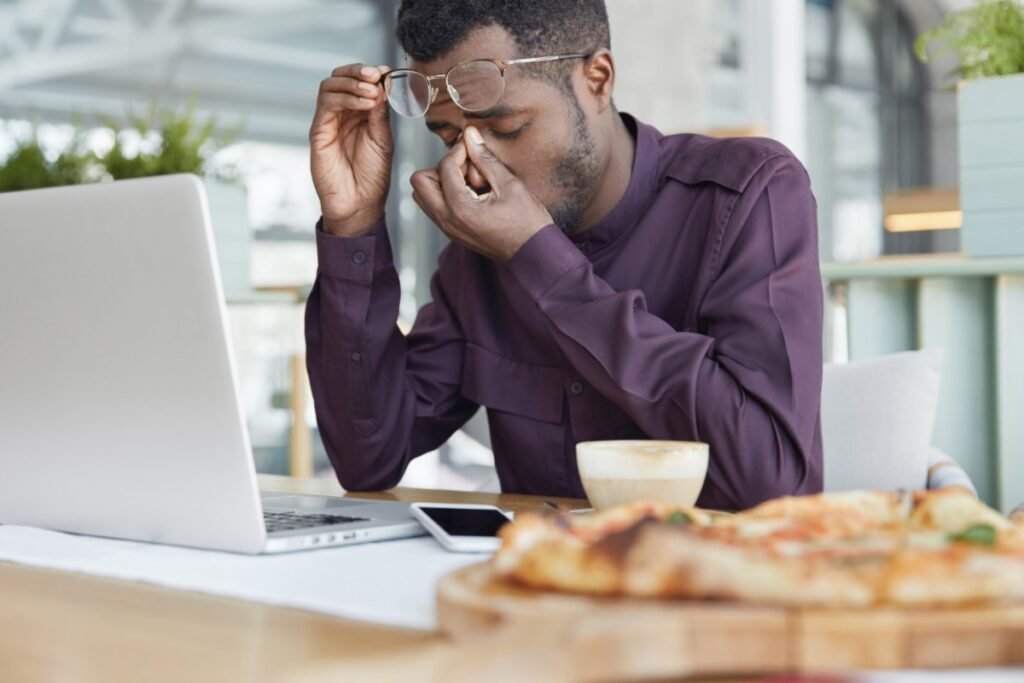 La imagen muestra a un hombre negro, con una camisa formal color vinotinto, sentado frente a un escritorio de madera. Sostiene sus gafas con la mano derecha por encima de la frente. Con la mano izquierda, presiona la zona entre los ojos y la nariz, a modo de preocupación.