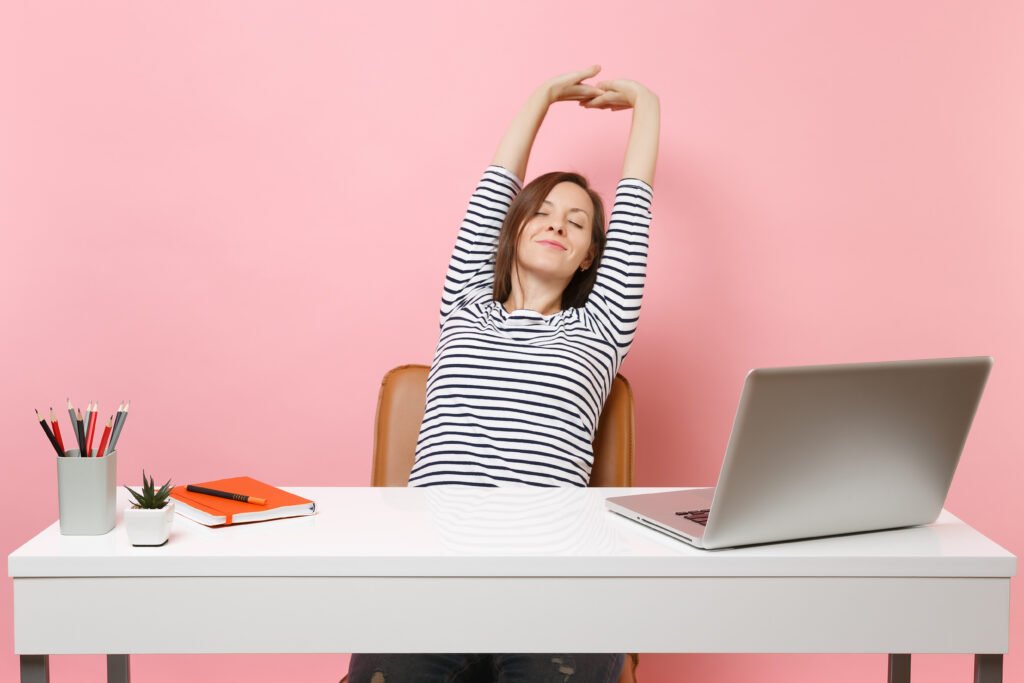 La imagen muestra a una mujer blanca con el pelo castaño liso sentada frente a un escritorio blanco, en el que hay una laptop plateada, un portalápices, una pequeña maceta y un cuaderno. La mujer tiene los brazos levantados por encima de la cabeza, como si se estuviera estirando. 