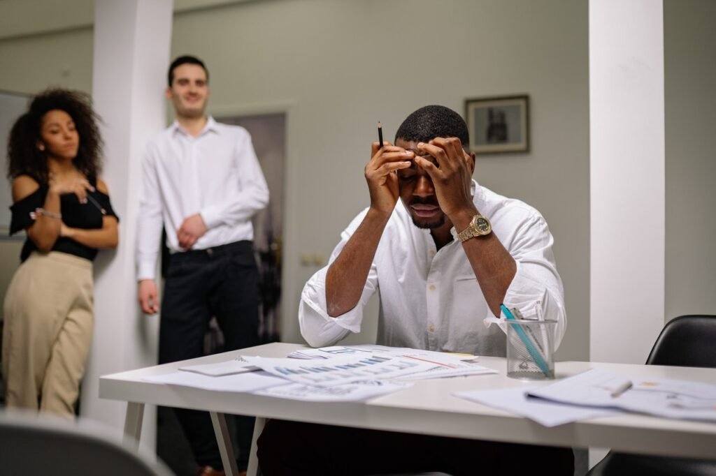 Um homem negro está sentado em frente a uma mesa de escritório, está com as mãos na cabeça e seu rosto apresenta uma expressão triste. Ao fundo há uma mulher negra e um homem branco, que olham para o homem negro.  