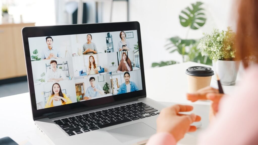La imagen muestra una laptop plateada sobre una mesa blanca. En la pantalla se puede ver una videollamada con nueve participantes. En la esquina derecha de la imagen, ligeramente borrosa, hay una mujer con una blusa rosa.