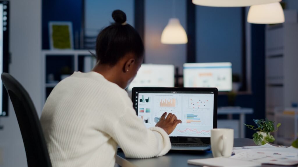 La imagen muestra a una mujer negra con cabello recogido en un moño, sentada de espaldas a la fotografía y delante de un escritorio. Está trabajando en una laptop negra y en la pantalla de esta se pueden ver unos gráficos de colores, como si fuera un informe de resultados.