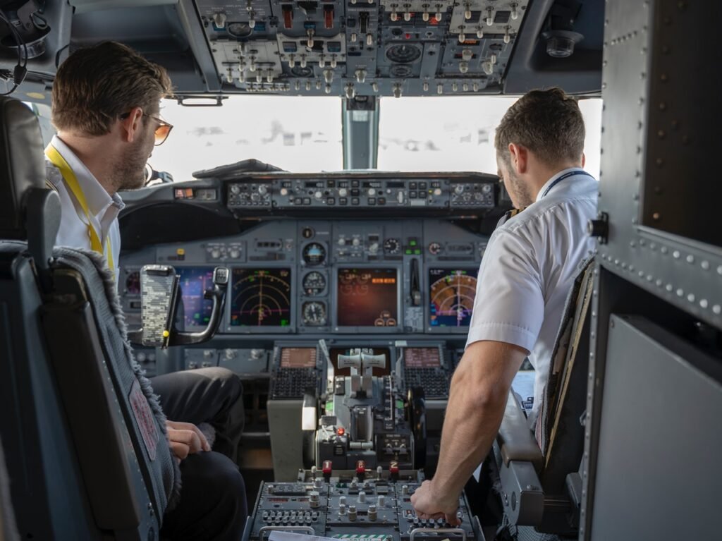 Dois homens brancos estão em uma cabine de uma aeronave e parecem ser os pilotos dela. Eles estão de costas para a imagem e é possível ver o painel do avião em frente a eles.
