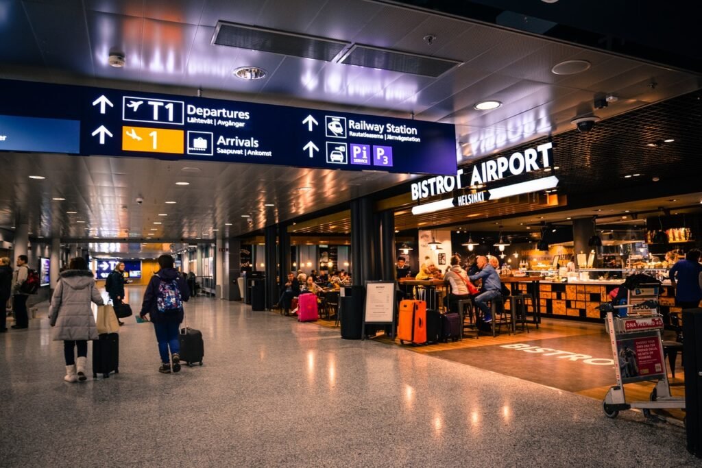 O saguão de um aeroporto aparece na imagem. Nele é possível ver algumas pessoas transitando com malas de viagem e um restaurante à direita.
