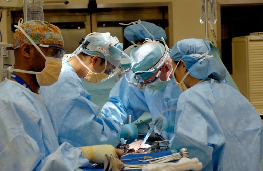 Uma equipe médica com quatro pessoas aparece em uma sala de cirurgia. Todos vestem um uniforme azul de tecido TNT, usam máscara e óculos de segurança.