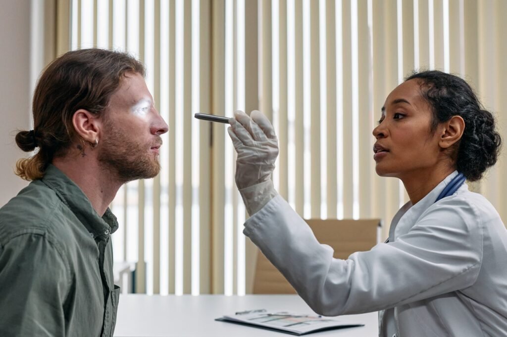  Um homem branco de cabelos castanhos-claros lisos presos está em frente a uma médica negra de cabelos pretos. Ela veste um jaleco branco e aponta uma pequena lanterna em direção ao olho do homem.