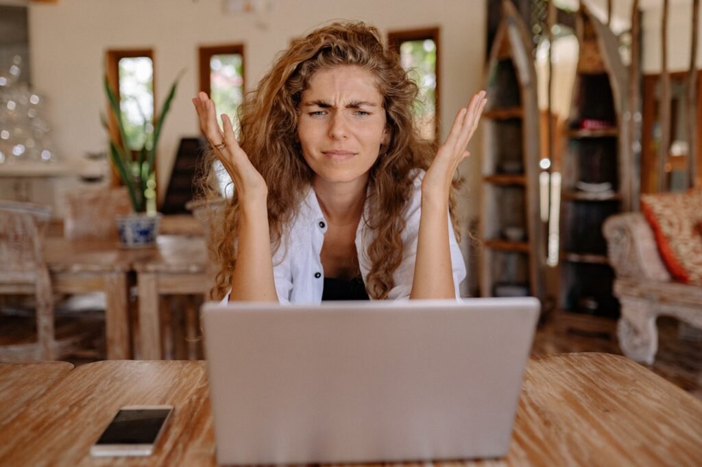La imagen muestra a una mujer blanca con cabello ondulado y castaño claro sentada frente a un escritorio de madera. Tiene las manos abiertas delante de la cara y está frunciendo ligeramente el ceño, como si se cuestionara lo que está viendo en la laptop que tiene frente a ella.