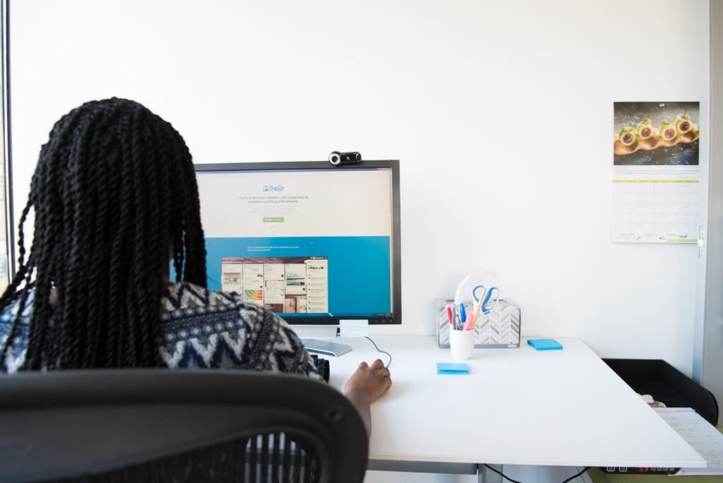 La imagen muestra a una mujer negra con el cabello trenzado sentada delante de una computadora. Está de espaldas a la imagen y su brazo derecho sostiene un mouse. En el ordenador se puede ver la pantalla de inicio de Trello.