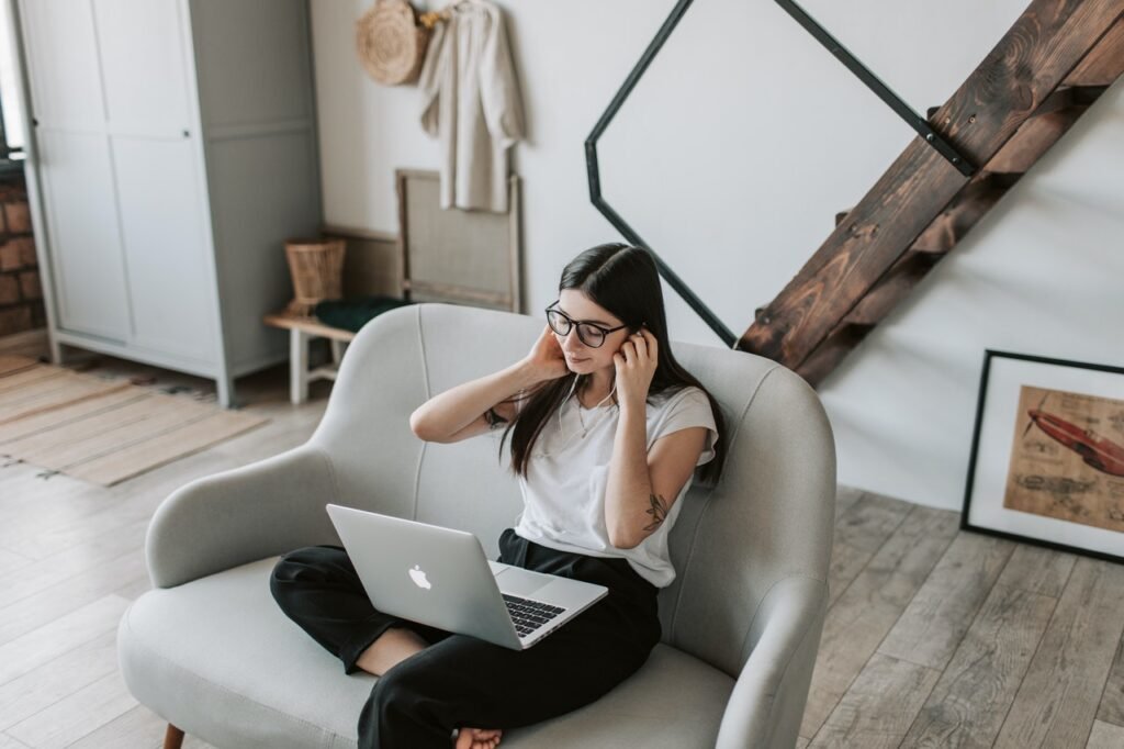 La imagen muestra a una mujer blanca con cabello castaño oscuro y liso que está sentada en un sillón gris. Ella está con una laptop plateada en su regazo y, con ambas manos, se está poniendo sus audífonos.