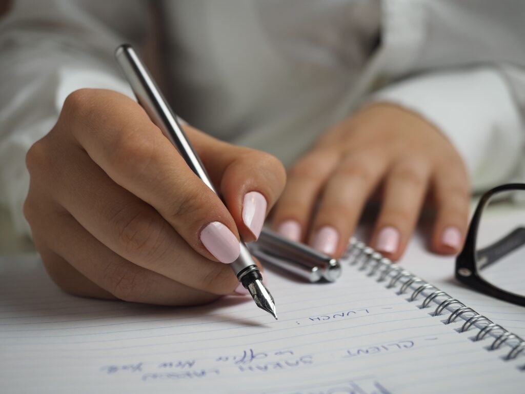 As mãos de uma pessoa negra, vestida com uma camiseta branca, aparecem na imagem. Ela está com uma caneta em mãos, fazendo anotações em um caderno. É possível ver outra caneta sobre o caderno e também óculos parcialmente aparentes.