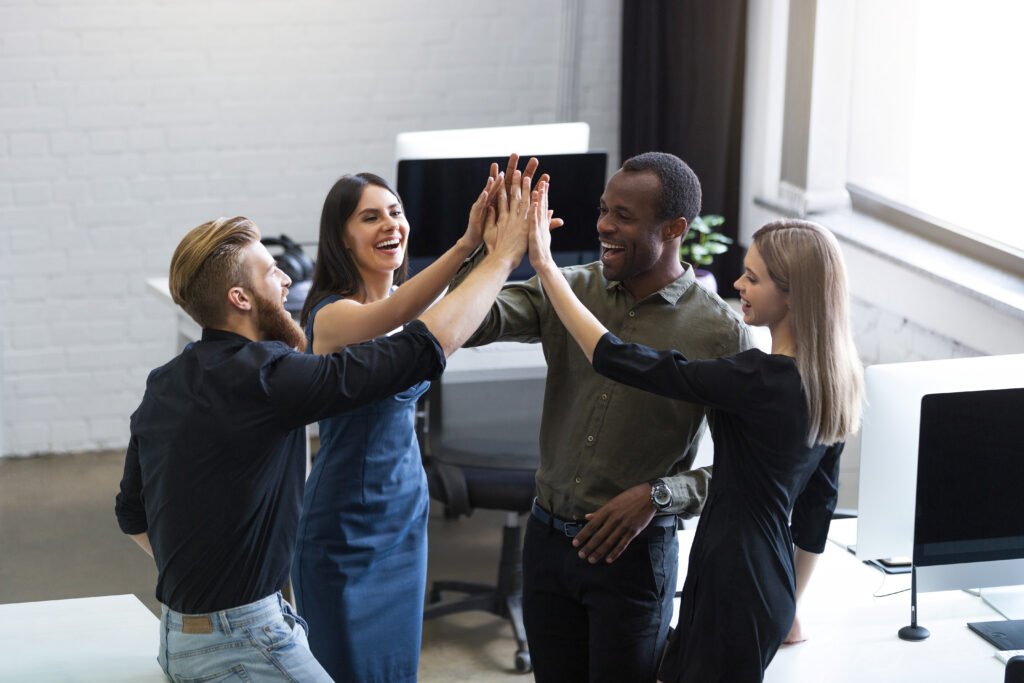 Em um escritório, duas mulheres brancas, uma loira e outra morena, e dois homens, um negro e um branco, estendem as mãos em um high five.