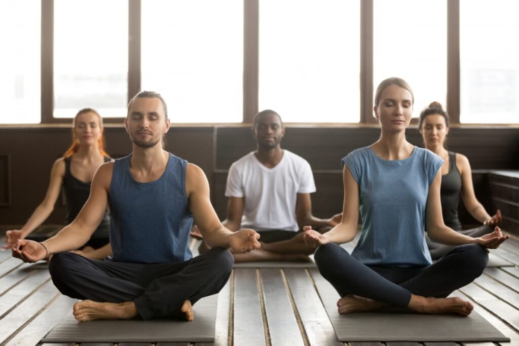 Un grupo de cinco personas están sentadas en posición de loto, con las piernas cruzadas y los brazos sobre las rodillas. Todos tienen los ojos cerrados y meditan.