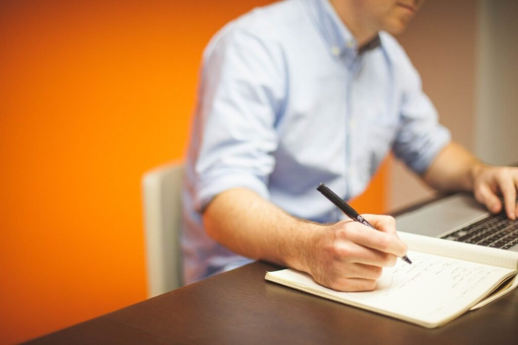 Um homem branco usando camisa azul sentado em frente a uma mesa com um laptop e um caderno onde anota algo.