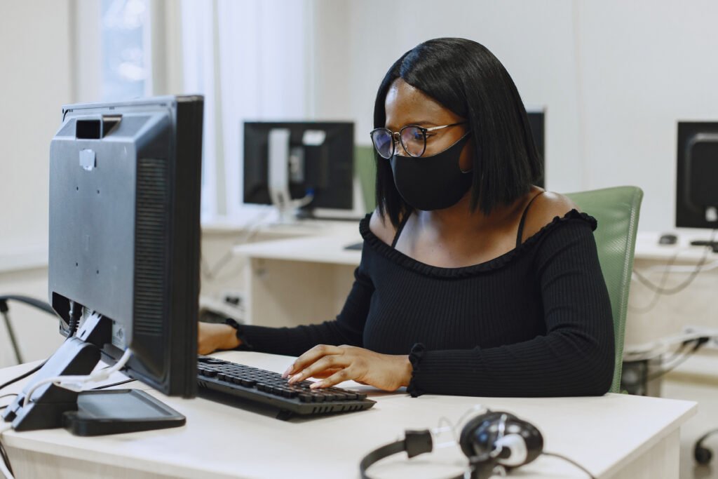 Mulher negra de cabelos pretos usando óculos, máscara e blusa também pretas, sentada em frente a um computador onde digita.