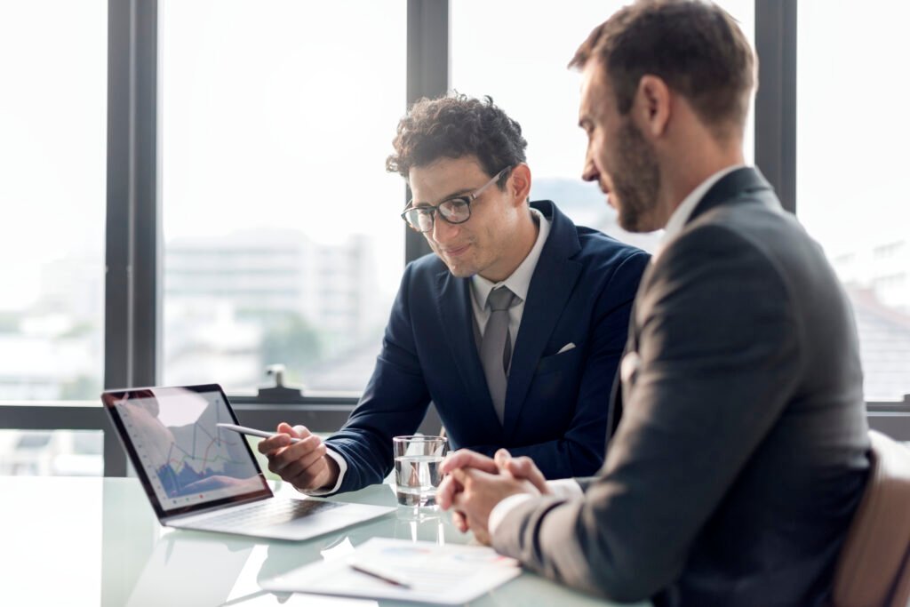 Dois homens brancos estão sentados em frente a uma mesa olhando um laptop no qual há alguns gráficos, representando uma avaliação de desempenho.